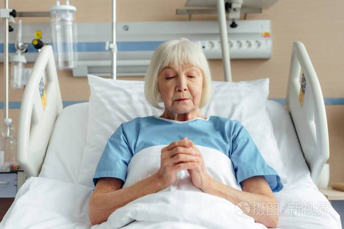 老年妇女双手交叉躺在床上在医院里祈祷